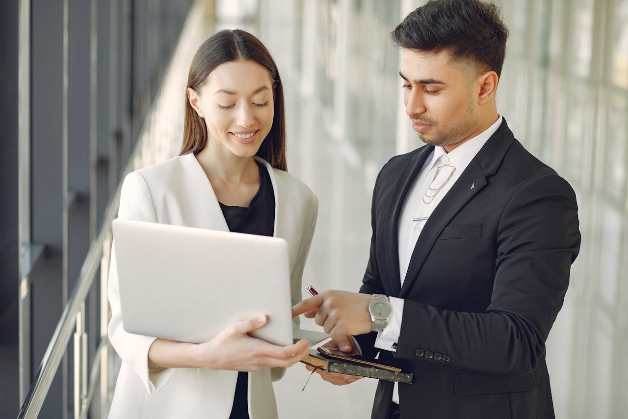 Concentrated diverse male and female employees in formal clothes standing in modern office corridor and browsing laptop while discussing business project together