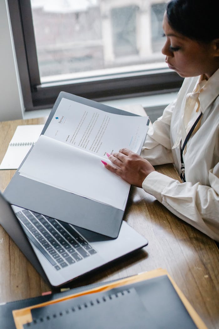 From above side view of crop African American female manager reading report of project while working with laptop in office
